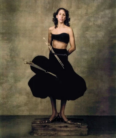 A fashion portrait of a slender woman wearing a black two-piece outfit with unique shoes, standing on a wooden crate against a painted backdrop.