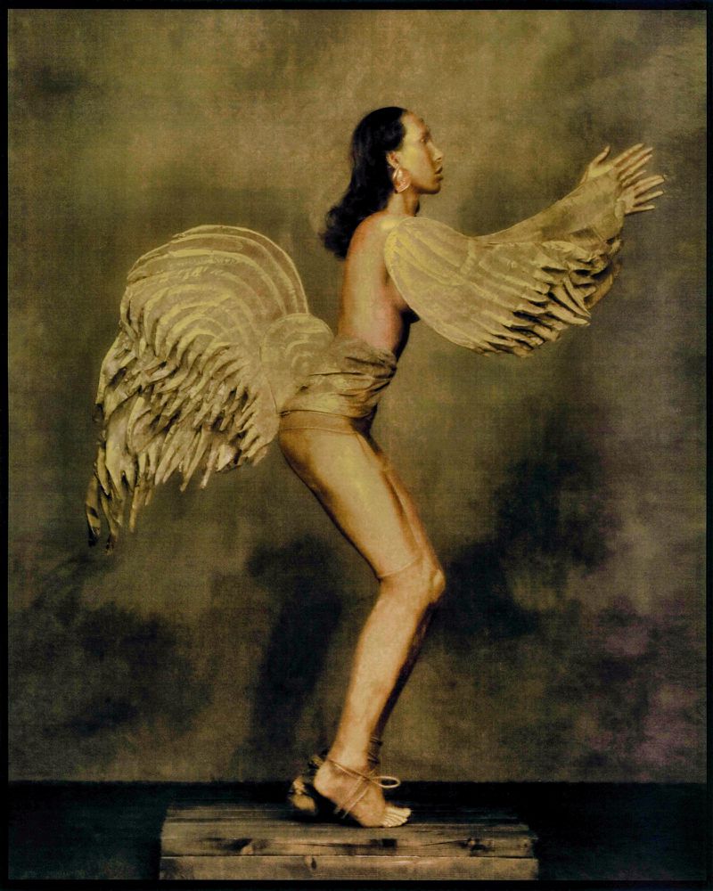 Surreal full body portrait of an androgynous person with outstretched arms, wearing light gold sculptural garment and minimalist heeled sandals.