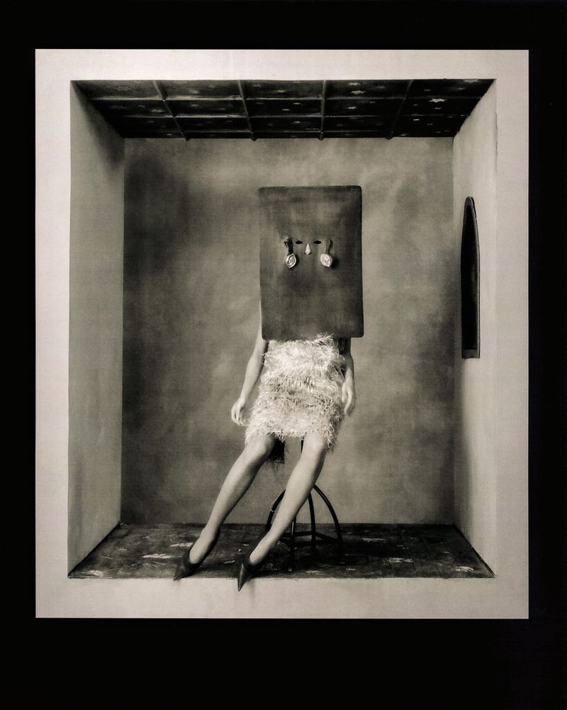 Monochrome photograph of a woman with a rectangular box over her head sitting on a stool indoors in front of a window.