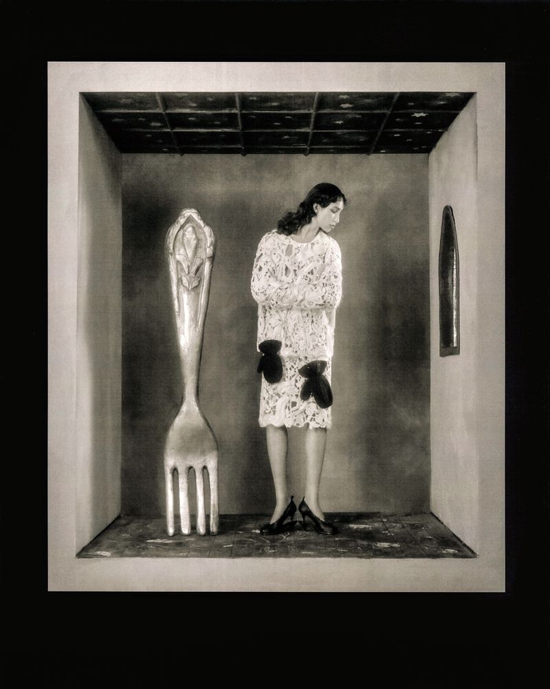 Monochrome photograph of a slender woman in a patterned white dress standing in a minimalist room with an oversized fork sculpture.