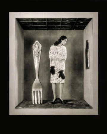 Monochrome photograph of a slender woman in a patterned white dress standing in a minimalist room with an oversized fork sculpture.