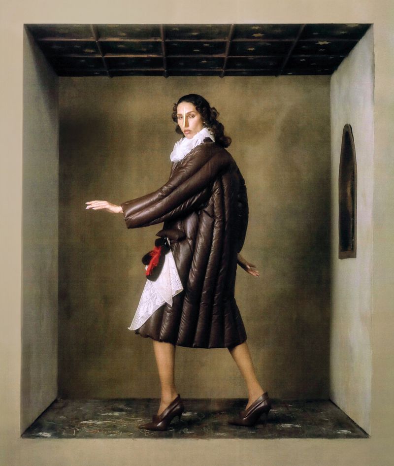 Fashion portrait of a woman in a brown coat with a white ruffled collar, posing in a three-walled studio space with a patterned ceiling.