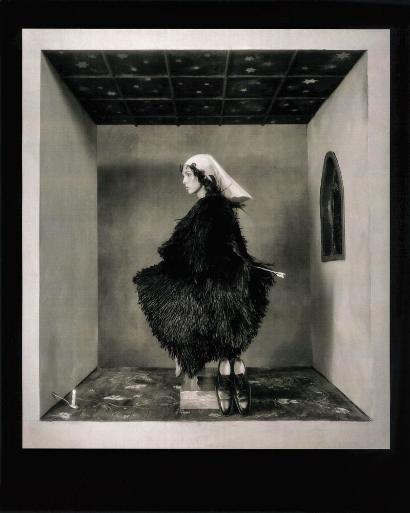 Artistic black-and-white studio shot of a woman in a feathered dress and headscarf standing in a box-shaped room with dark floral ceiling.