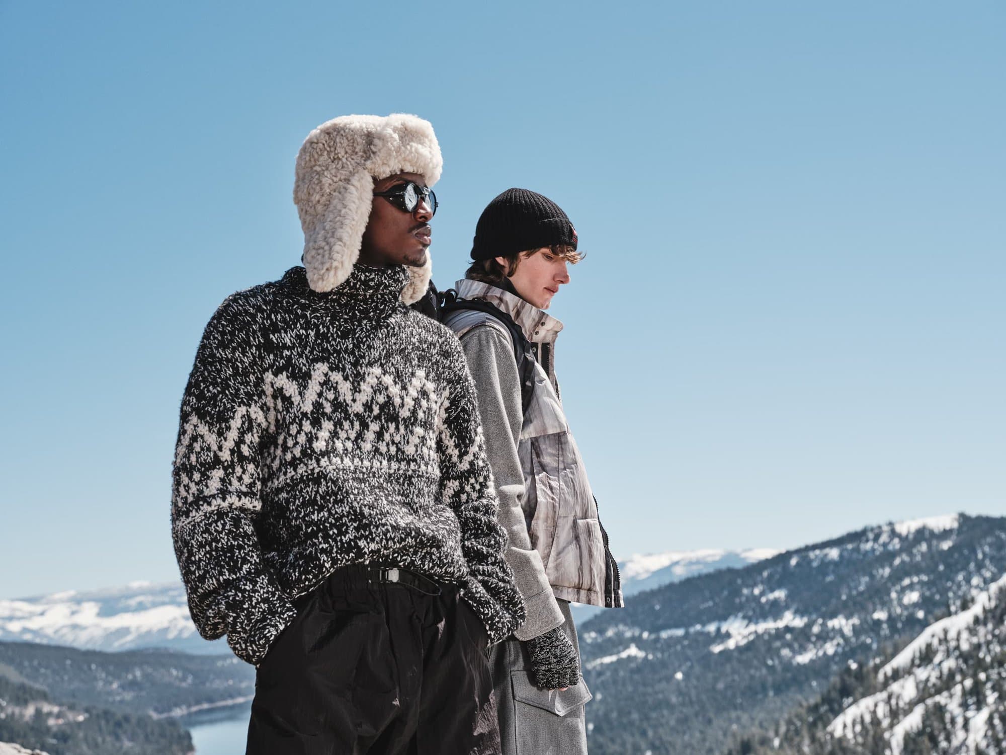 Two male models outdoors against a snowy mountain backdrop wearing winter apparel including a fluffy ushanka hat