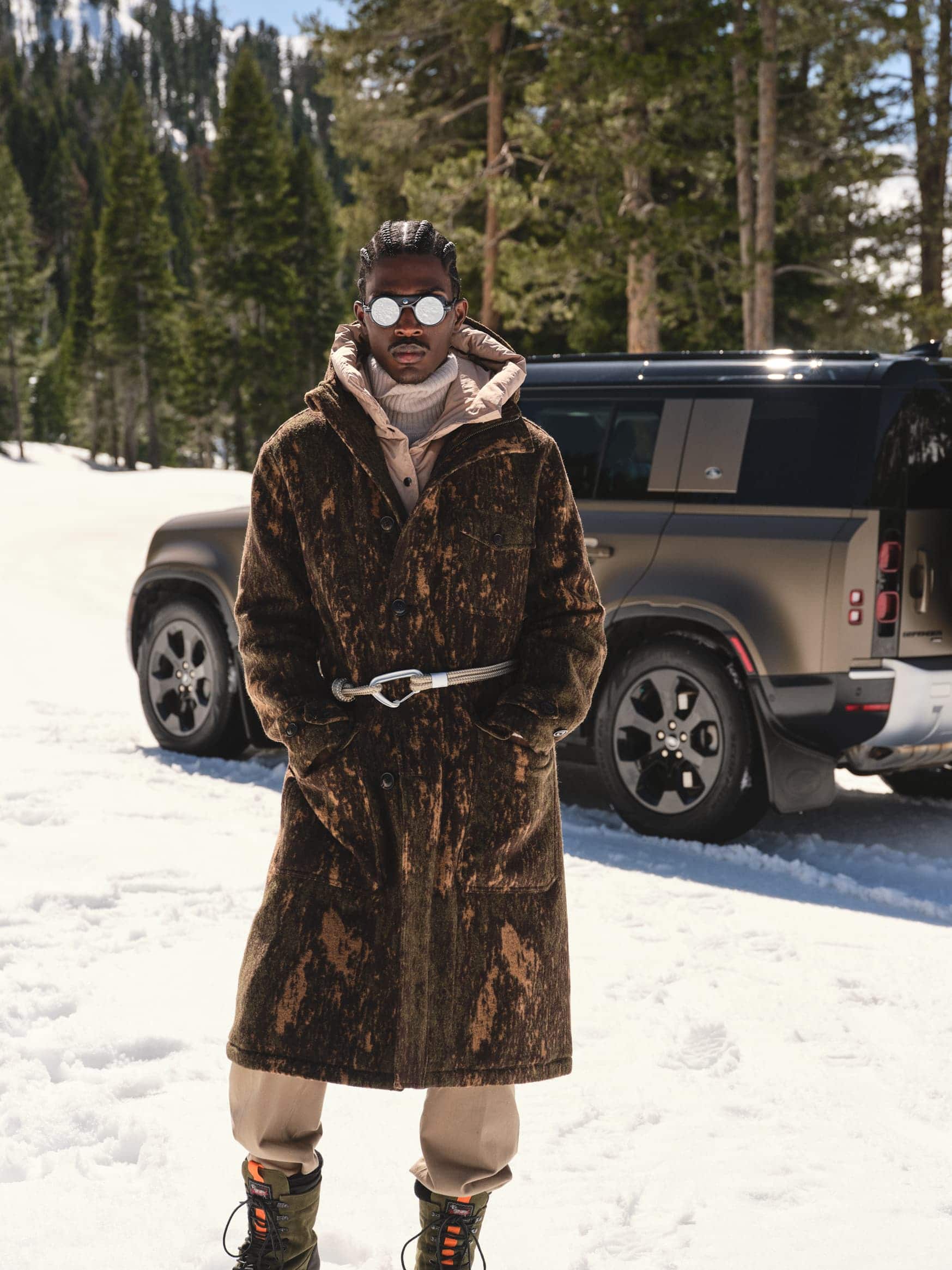 Man wearing a brown patterned coat and sunglasses standing in the snow in front of a dark SUV.