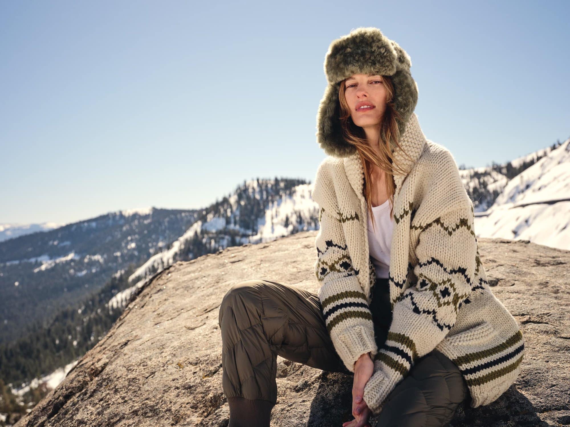 Model wearing a fuzzy green trapper hat and patterned cream cardigan seated on a snowy mountain ridge under a clear sky.