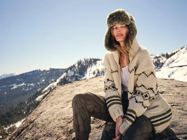 Model wearing a fuzzy green trapper hat and patterned cream cardigan seated on a snowy mountain ridge under a clear sky.