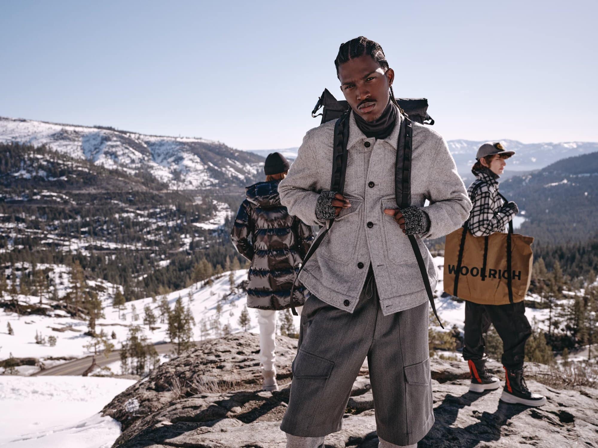 Three models wearing winter apparel outdoors on a rocky mountain ridge with snow-covered terrain and pine trees in the background.