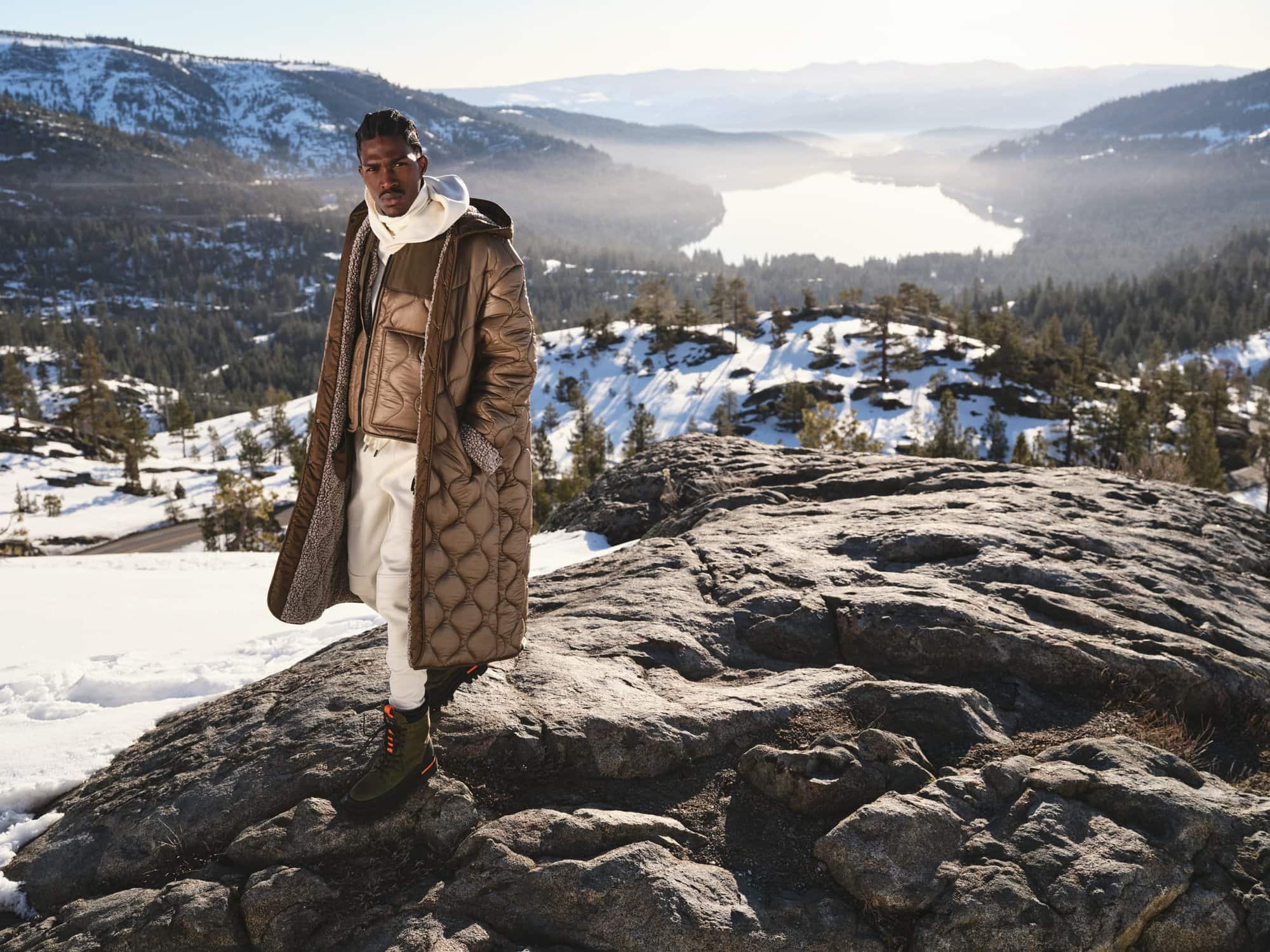 Model standing on snowy rock outcropping wearing long bronze quilted coat with lake and mountain view behind