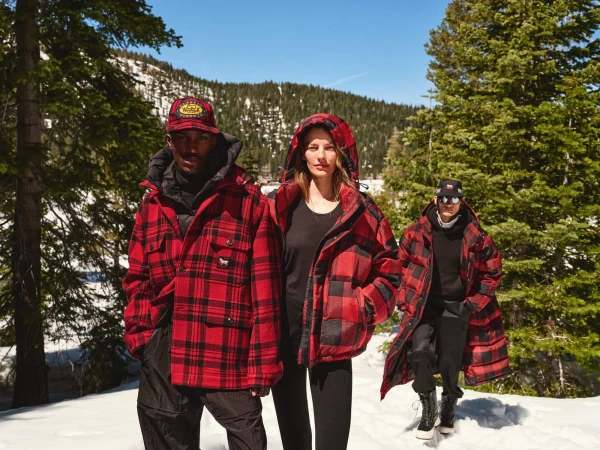 Three models wearing red and black buffalo plaid outerwear posing outdoors in a snowy mountain forest.
