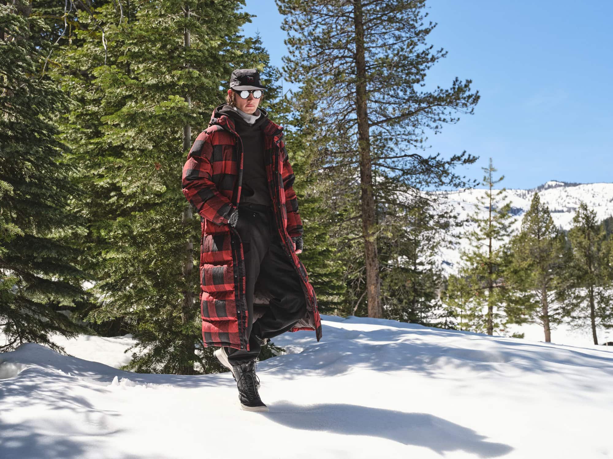 Model walking on a snow-covered path through a forest wearing a red and black plaid puffer coat and sunglasses.