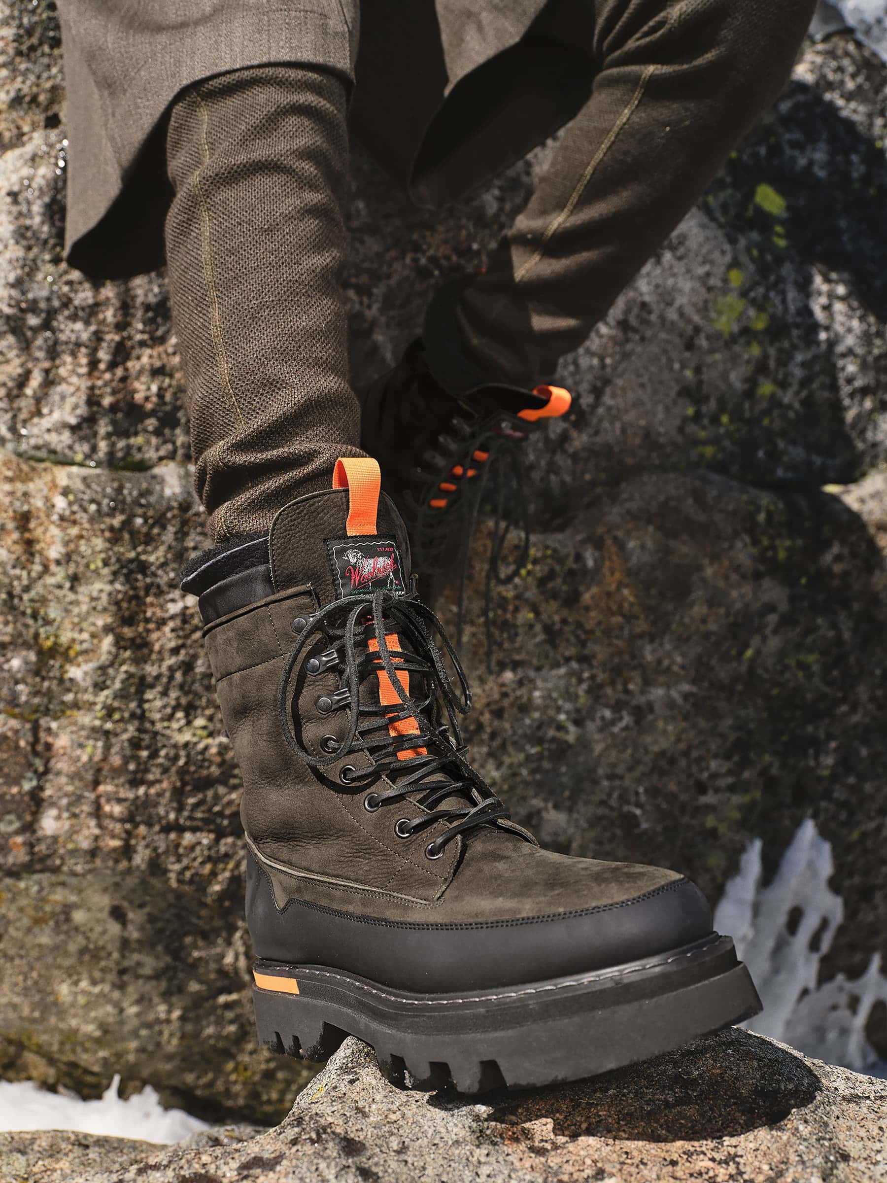 Close-up of rugged lace-up boot with thick black lug sole and orange pull tab worn on rocky terrain.