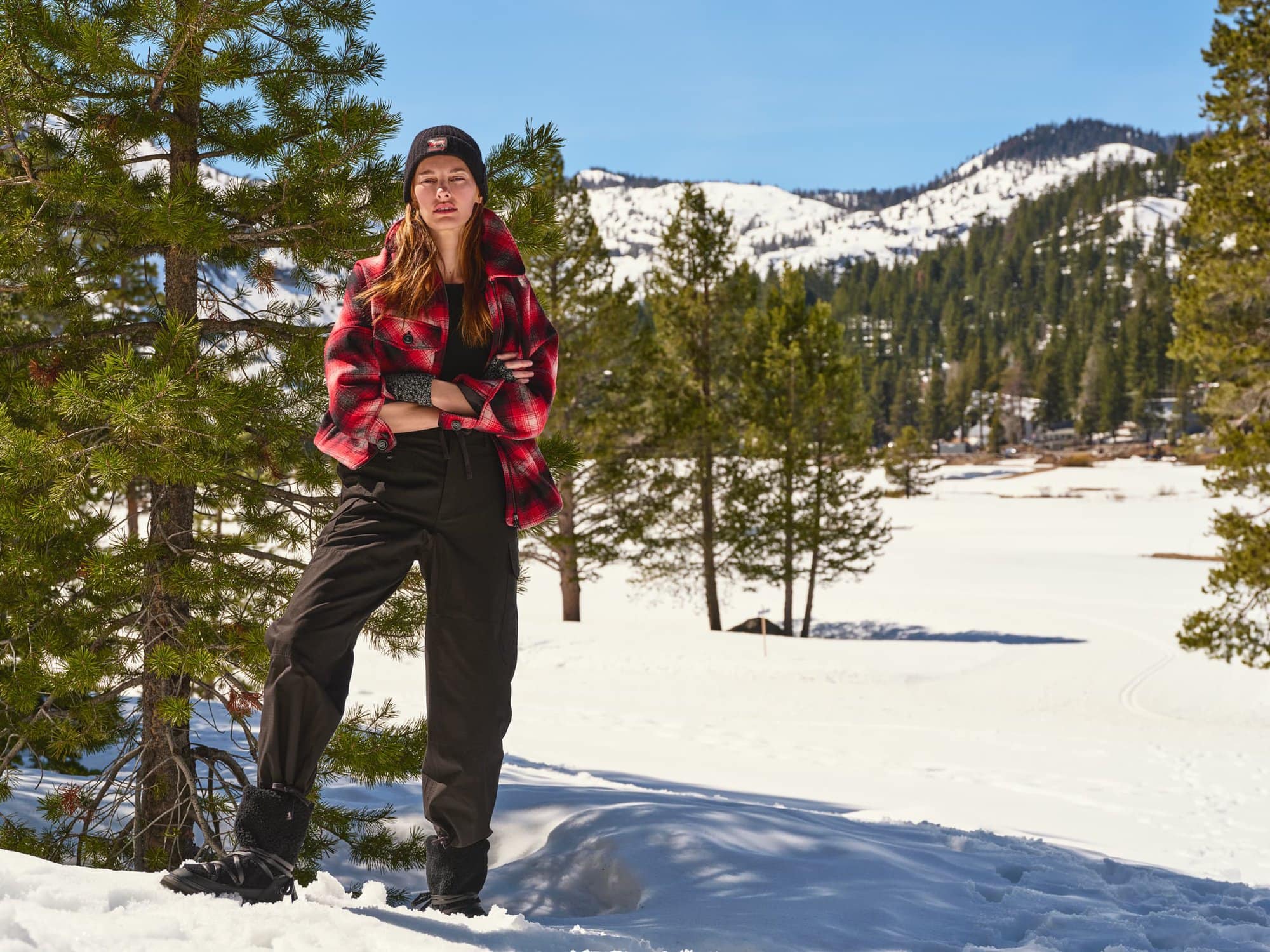 Model standing outdoors in a snowy landscape wearing a red plaid jacket, black beanie, and dark trousers.