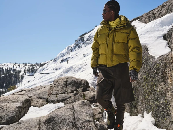 Man wearing yellow puffer jacket and cargo shorts standing on a snowy mountain ridge under a clear blue sky.