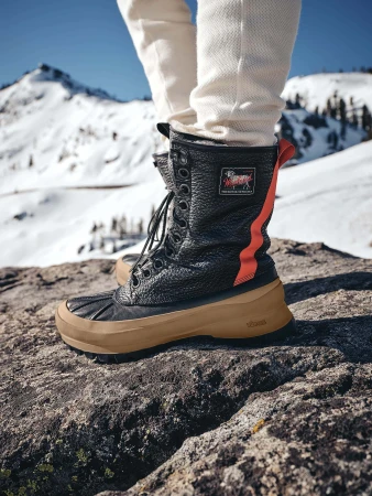 Close-up shot of black leather winter boots with brown rubber soles and white trousers standing on a snowy rocky mountain peak.