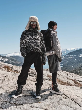Two models wearing winter outdoor apparel standing on a rocky mountain terrain under a clear blue sky