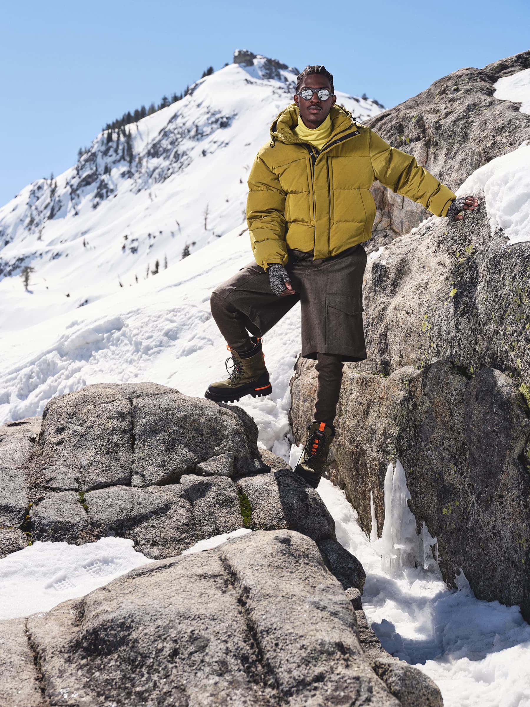 Man wearing a yellow puffer jacket standing on a snowy mountain rocky terrain under clear blue sky.