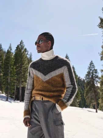 Man wearing a textured chevron sweater and sunglasses standing outdoors in a snowy mountain forest.