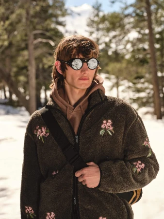 Model wearing round glacier sunglasses and an embroidered fleece jacket standing outdoors against a snowy forest