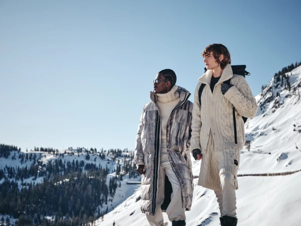 Two male models standing on a snowy mountain ridge wearing chunky knit sweaters and winter outerwear under a clear blue sky.