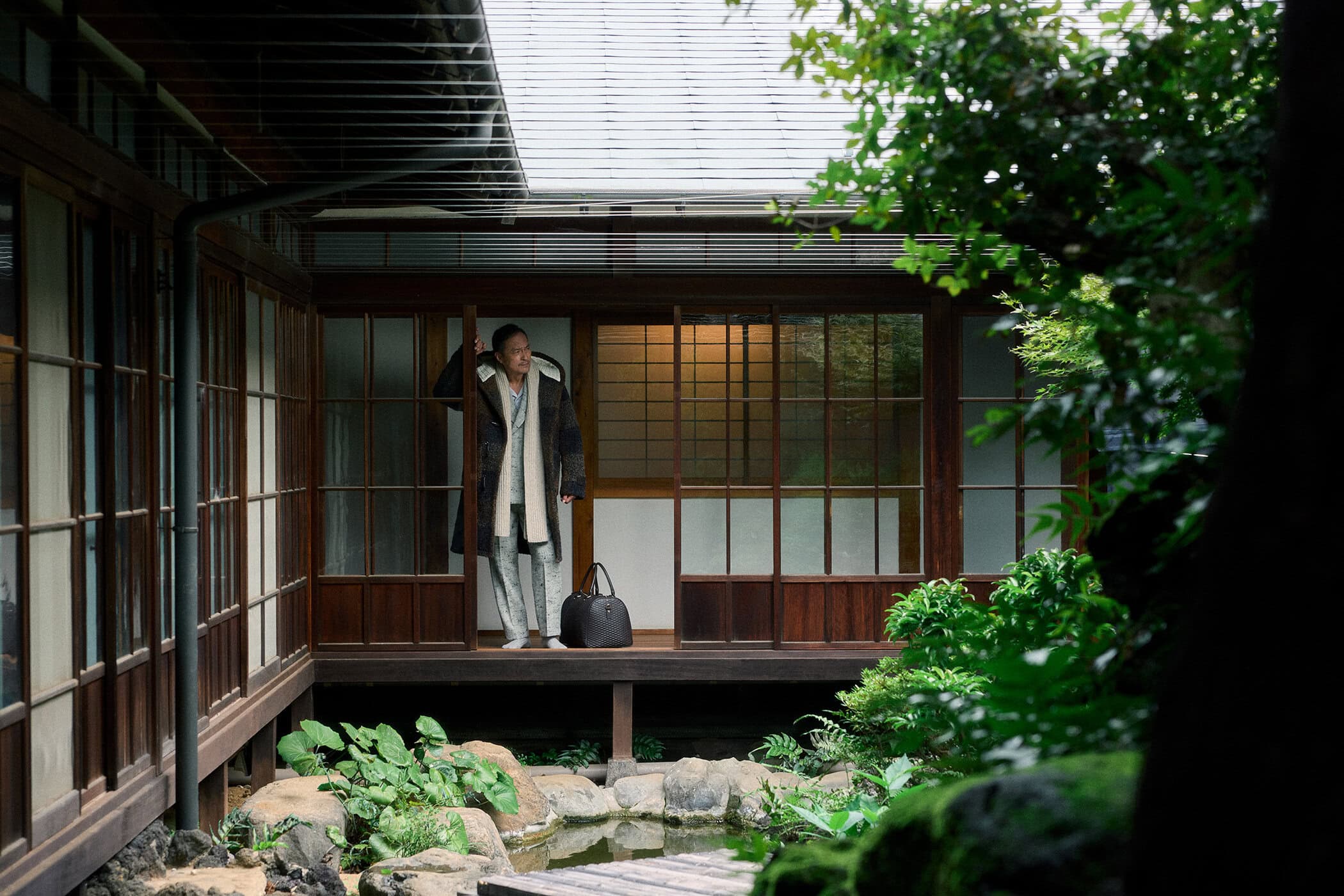 Ken Watanabe wearing a patterned coat and scarf standing in a traditional Japanese building with a courtyard garden