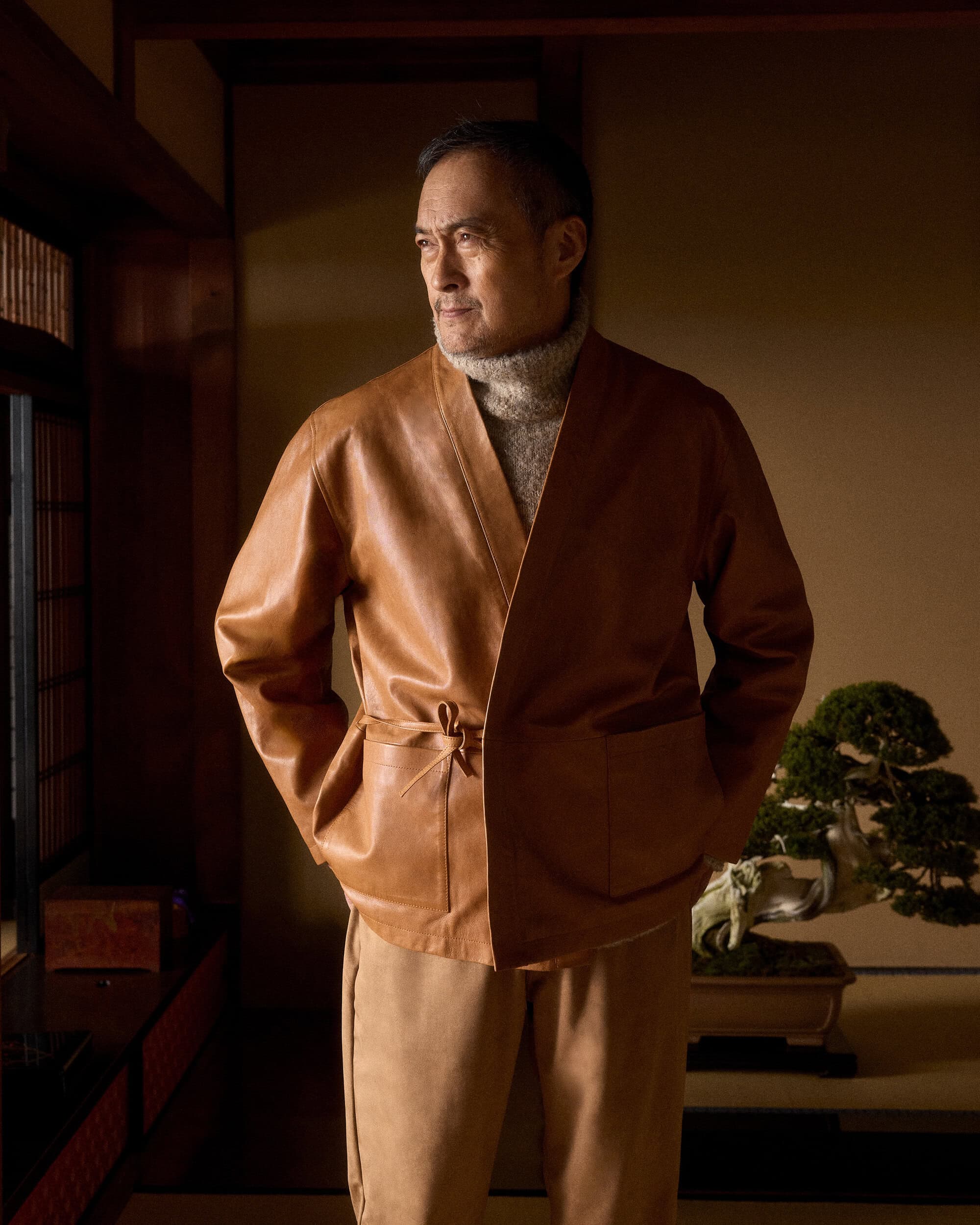 Ken Watanabe wearing a camel leather jacket and turtleneck inside a traditional Japanese room with a bonsai tree.