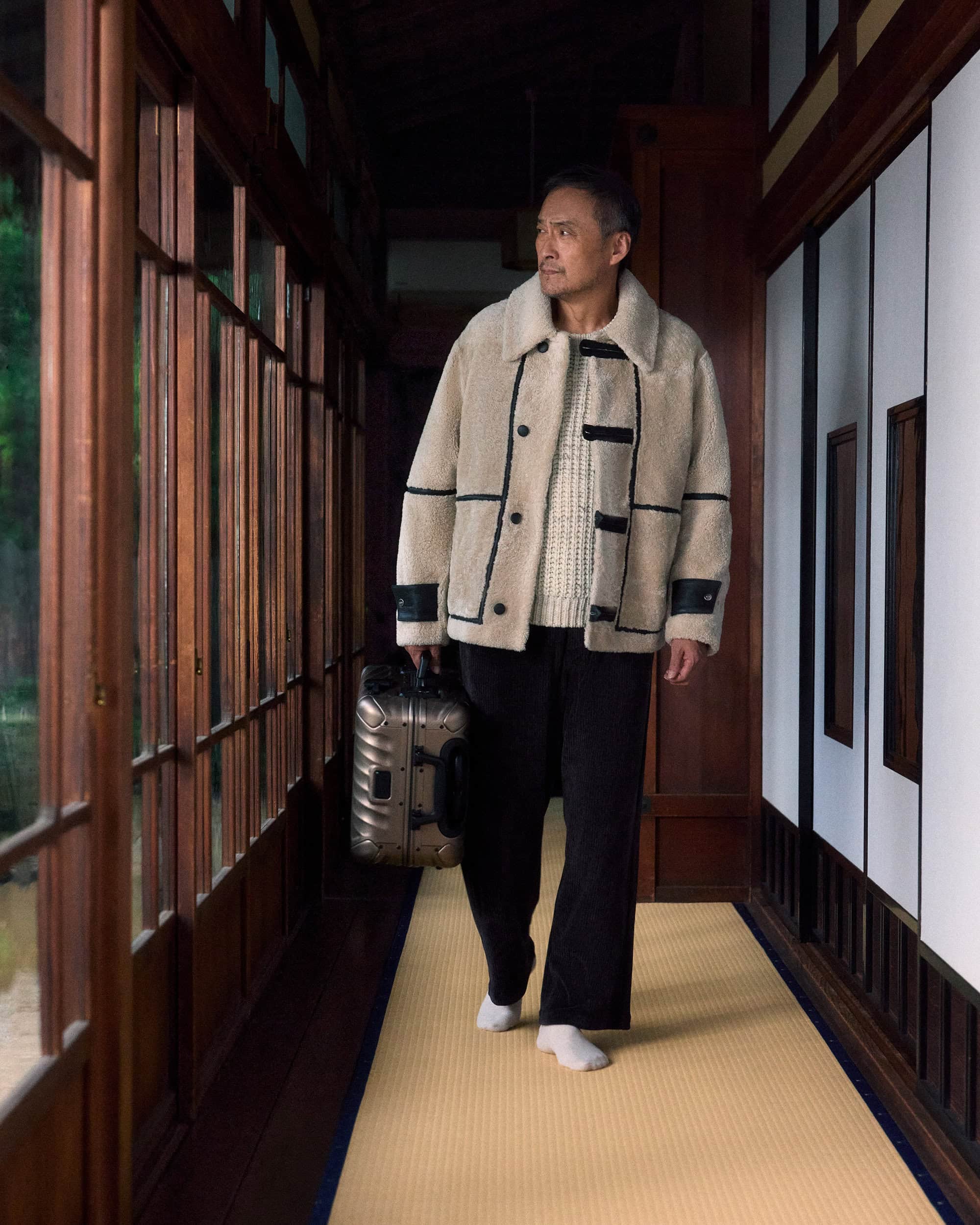 Ken Watanabe in a cream shearling jacket and dark trousers holding a silver suitcase in a traditional hallway.
