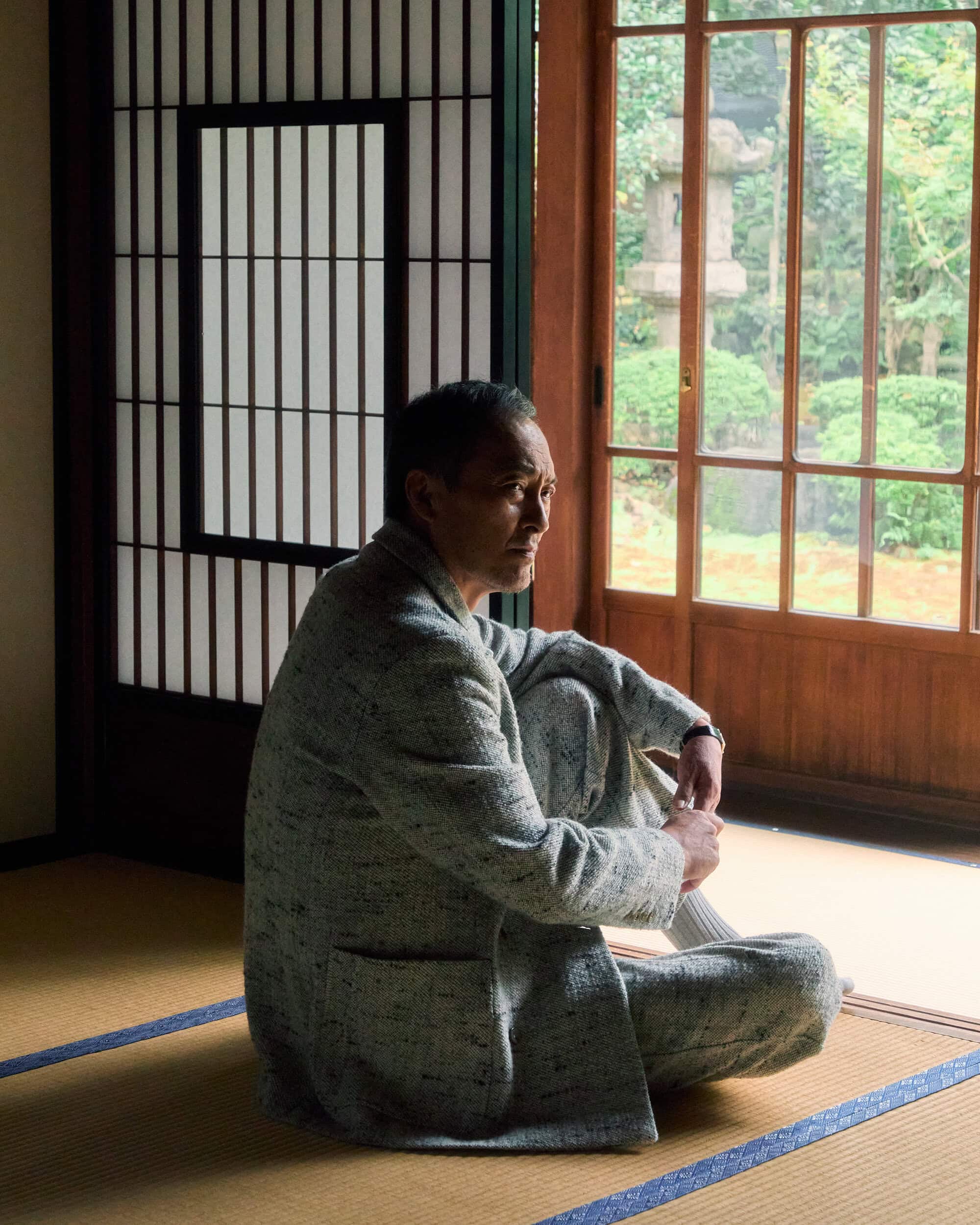 Ken Watanabe seated on tatami mats wearing a grey tweed patterned jacket near a traditional sliding door