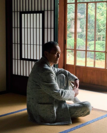 Ken Watanabe seated on tatami mats wearing a grey tweed patterned jacket near a traditional sliding door