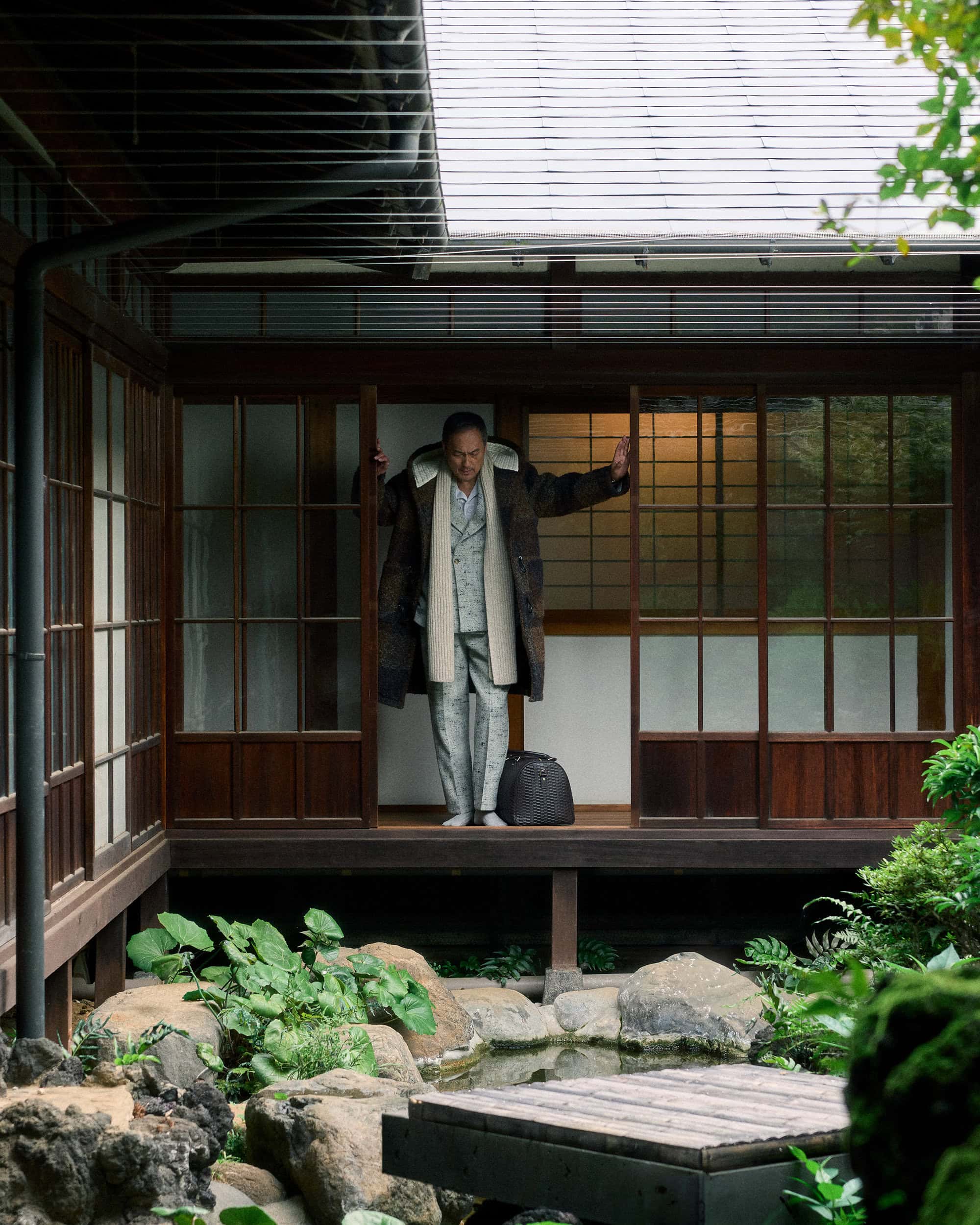 Ken Watanabe standing in a traditional Japanese wooden porch wearing a patterned suit and coat with a dark duffel bag