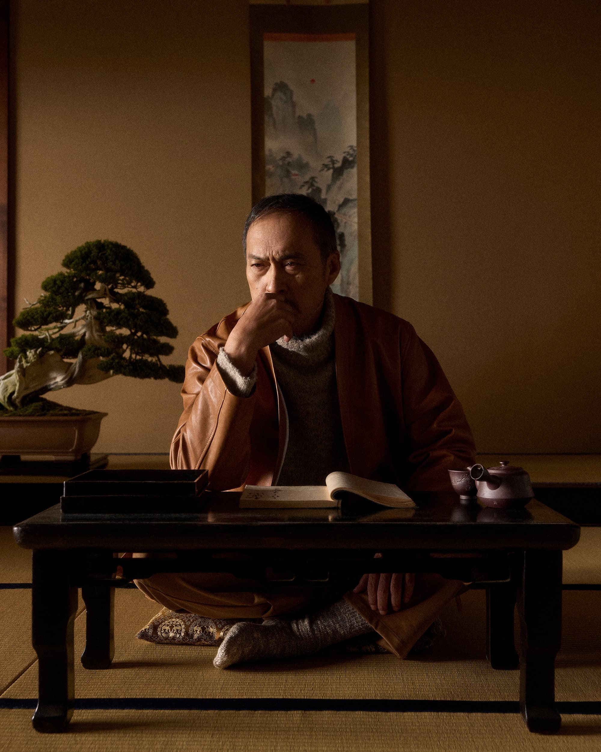 Actor Ken Watanabe sitting cross-legged at a low wooden table indoors, wearing a brown jacket and grey turtleneck sweater.