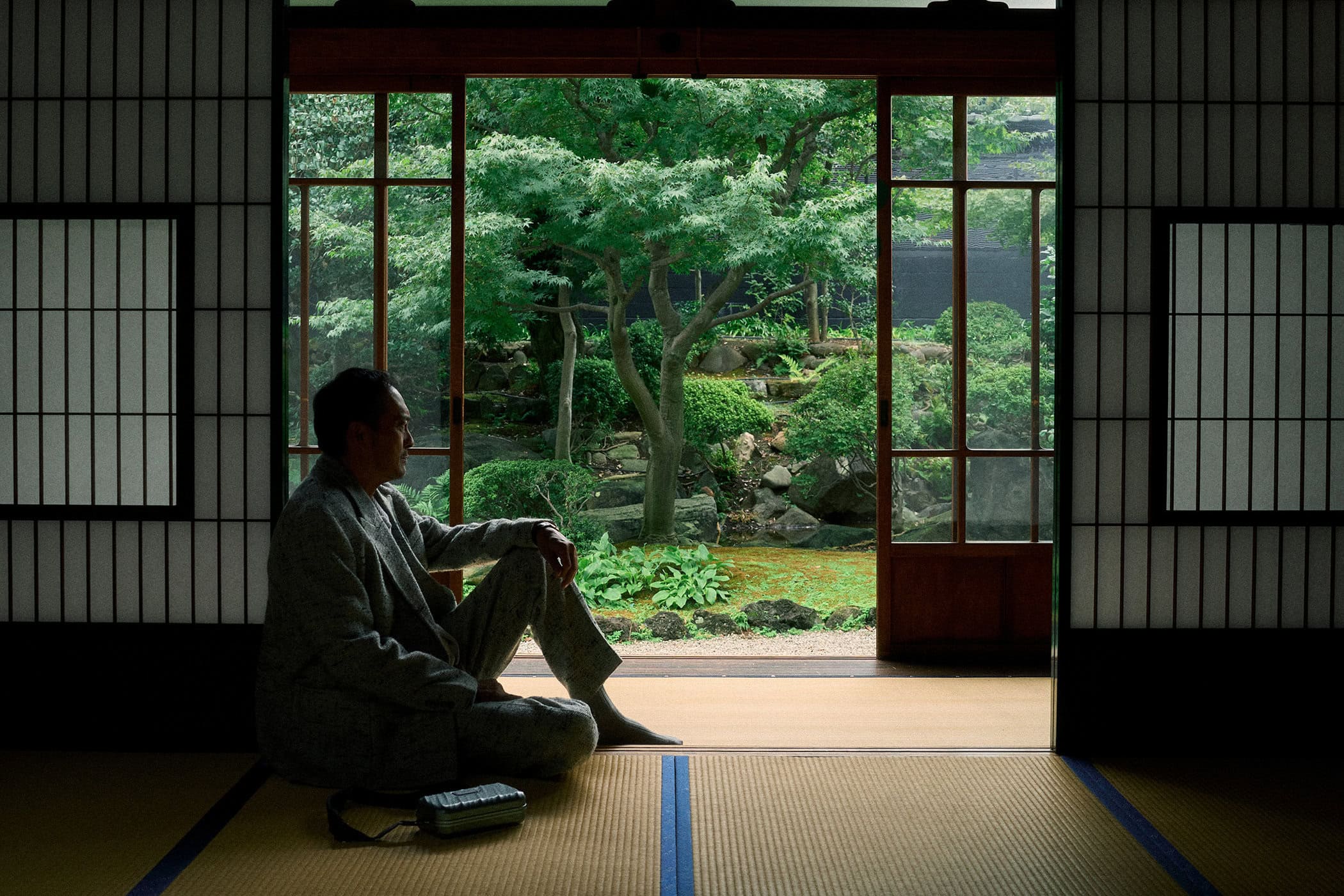 Ken Watanabe seated on tatami flooring inside a traditional Japanese room looking out at a lush garden.