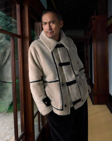 Ken Watanabe wearing a cream shearling coat leaning against a wood-framed window in an interior hallway for Kith