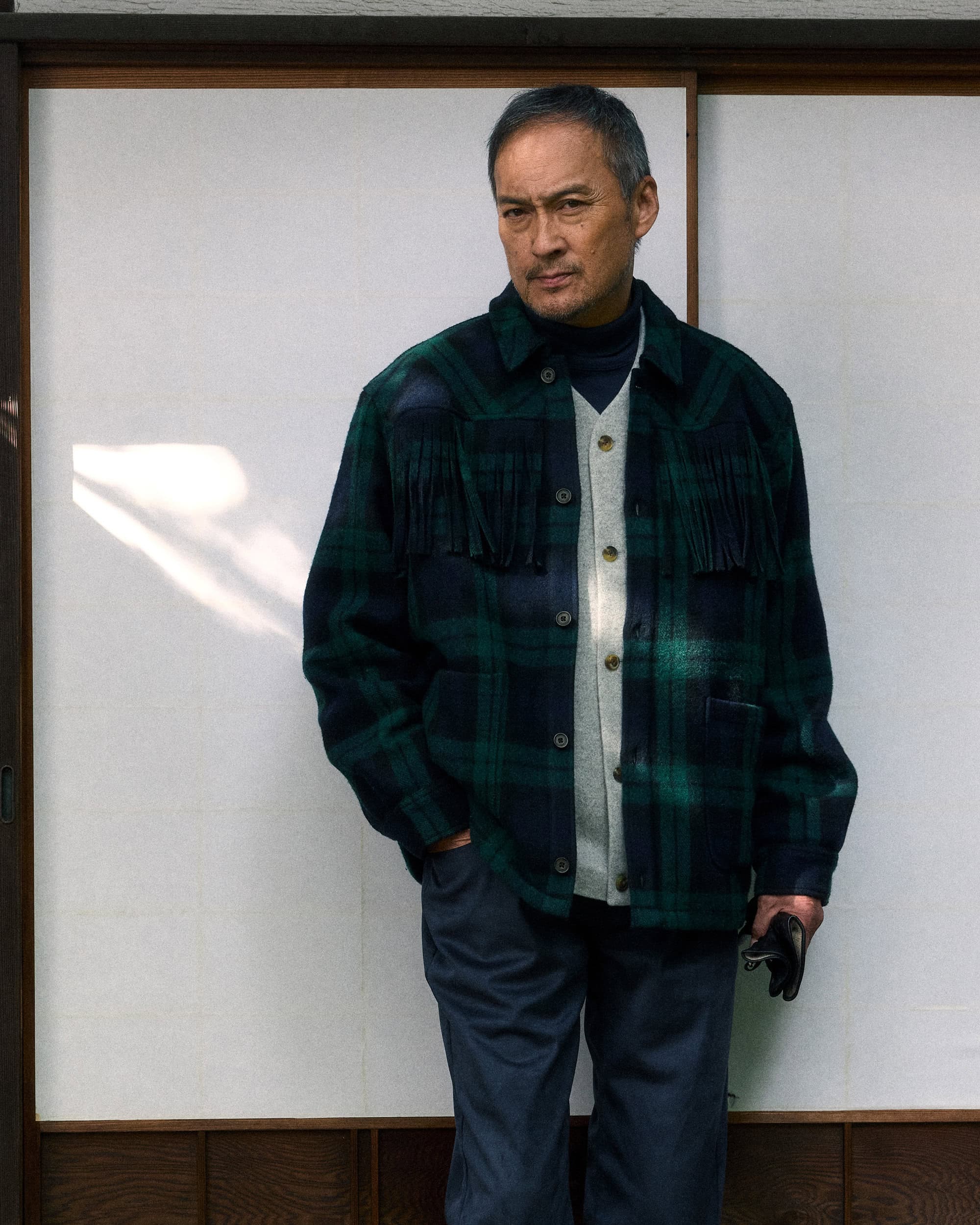 Ken Watanabe wearing a green plaid fringe jacket and dark trousers standing against a traditional shoji screen wall.