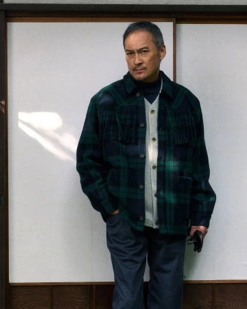 Ken Watanabe wearing a green plaid fringe jacket and dark trousers standing against a traditional shoji screen wall.