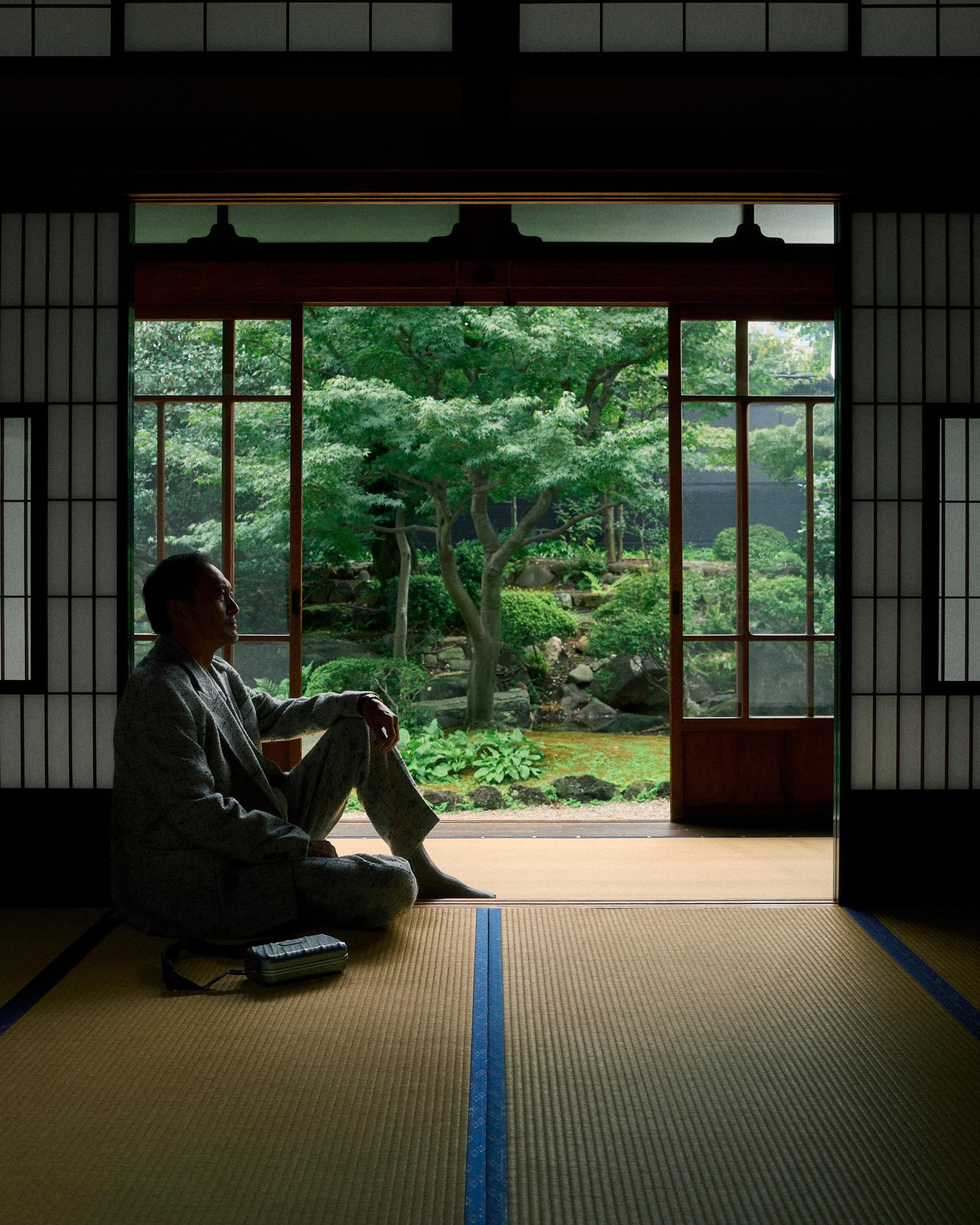 Ken Watanabe seated on tatami mats inside a traditional Japanese room looking out at a lush garden.