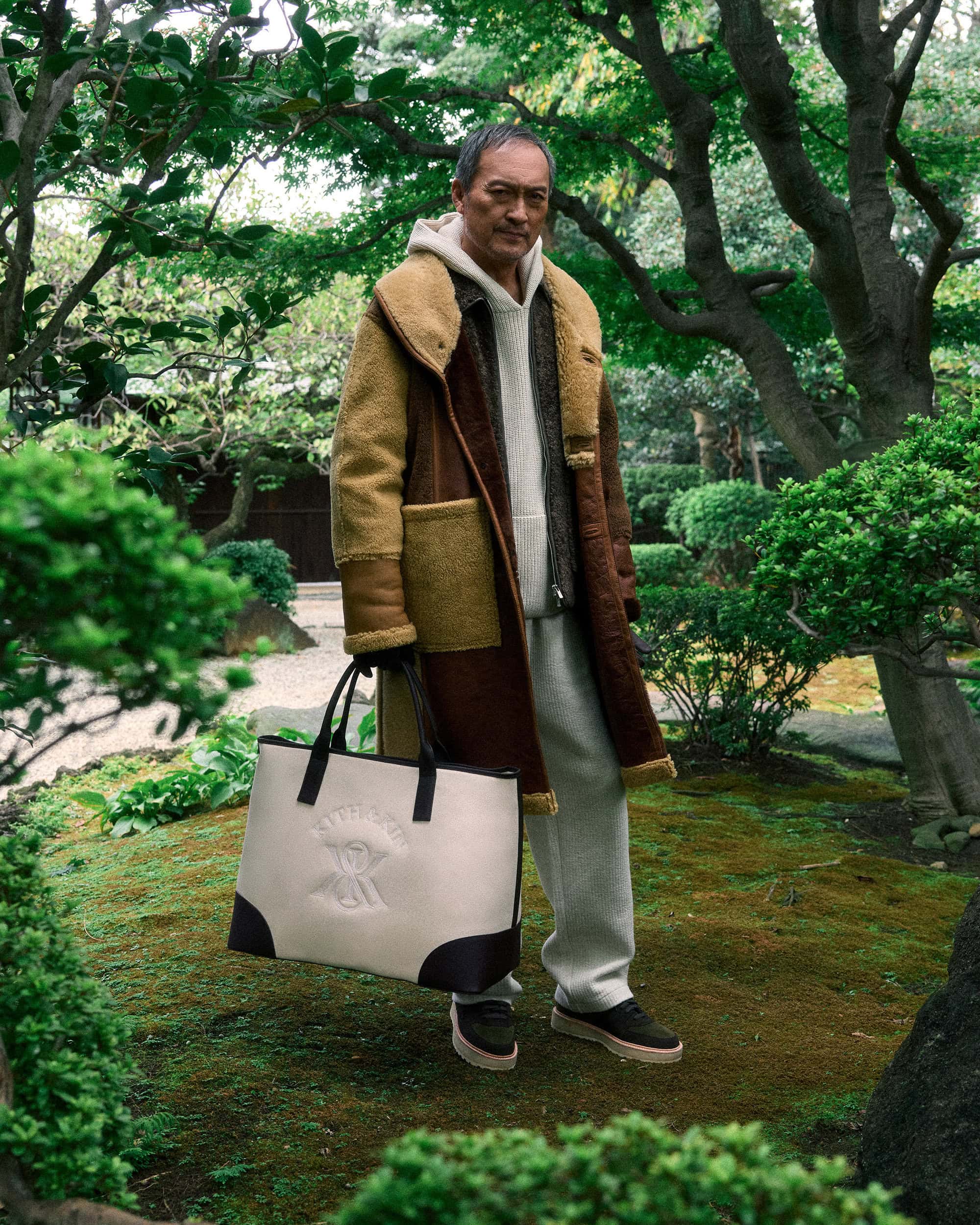 Ken Watanabe holding a large canvas tote bag wearing a shearling coat in a Japanese garden for Kith Winter 2024
