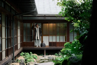 Ken Watanabe wearing a patterned coat and scarf standing in a traditional Japanese building with a courtyard garden