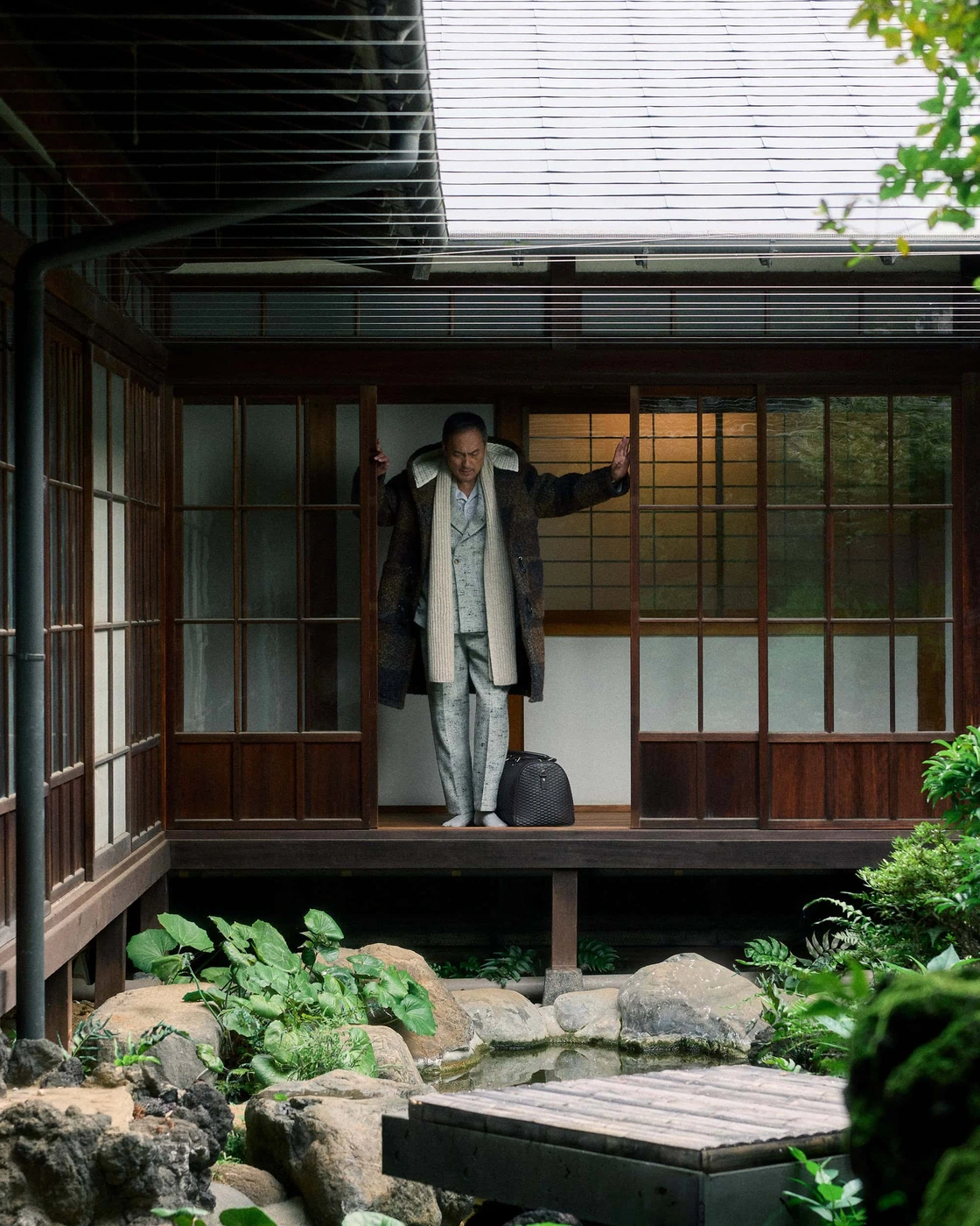 Ken Watanabe standing in a traditional Japanese wooden porch wearing a patterned suit and coat with a dark duffel bag