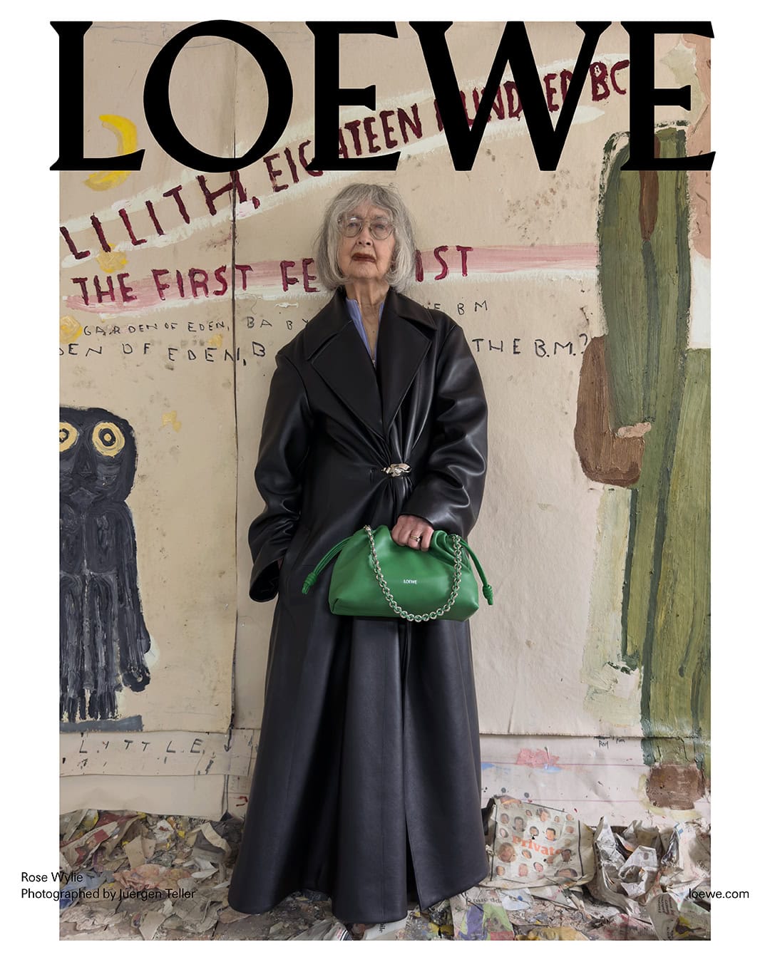 Elderly woman wearing a long dark leather coat and holding a green handbag in front of a painted backdrop