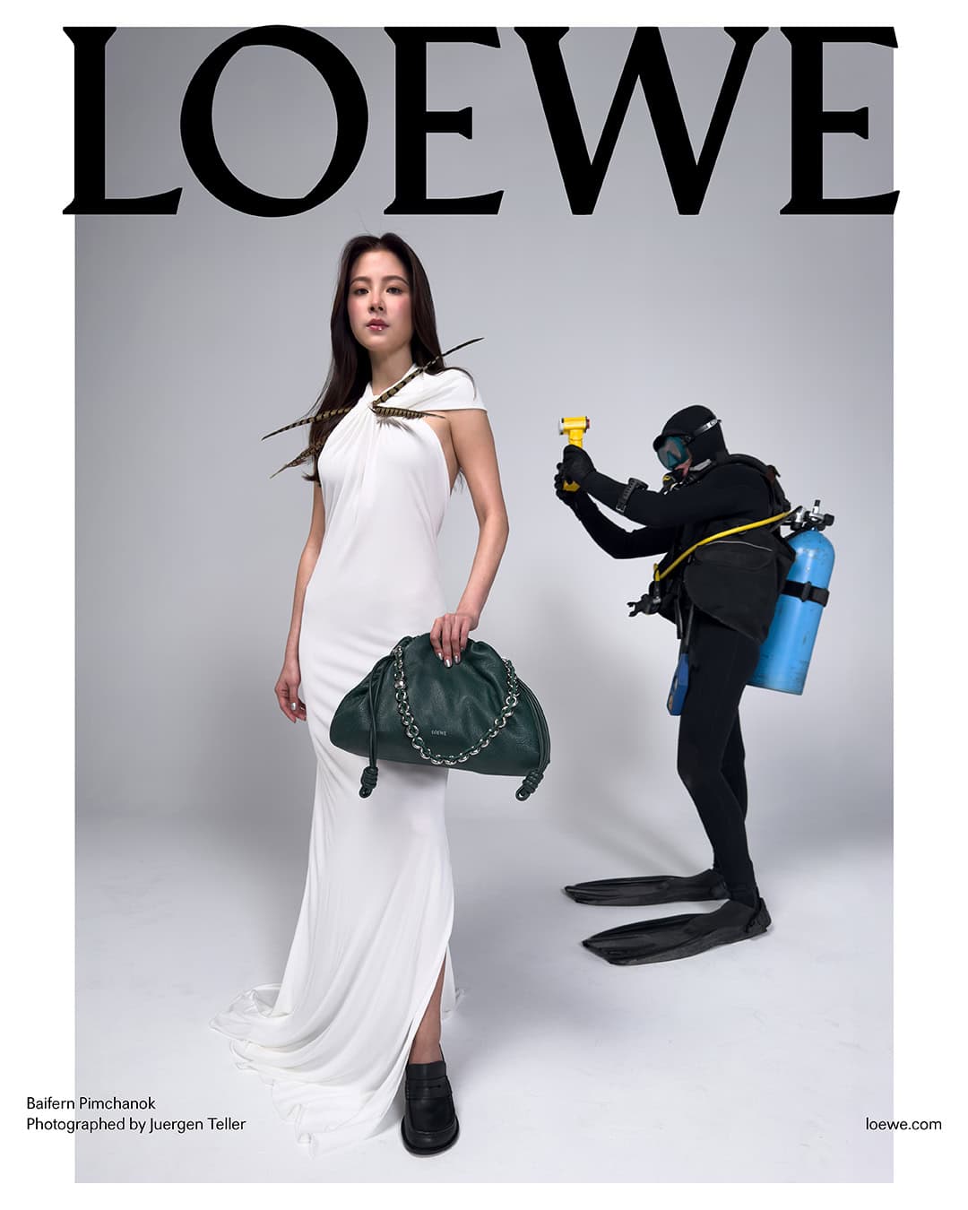 Baifern Pimchanok in a white halter dress holding a dark green handbag, with a diver standing behind her in a studio.