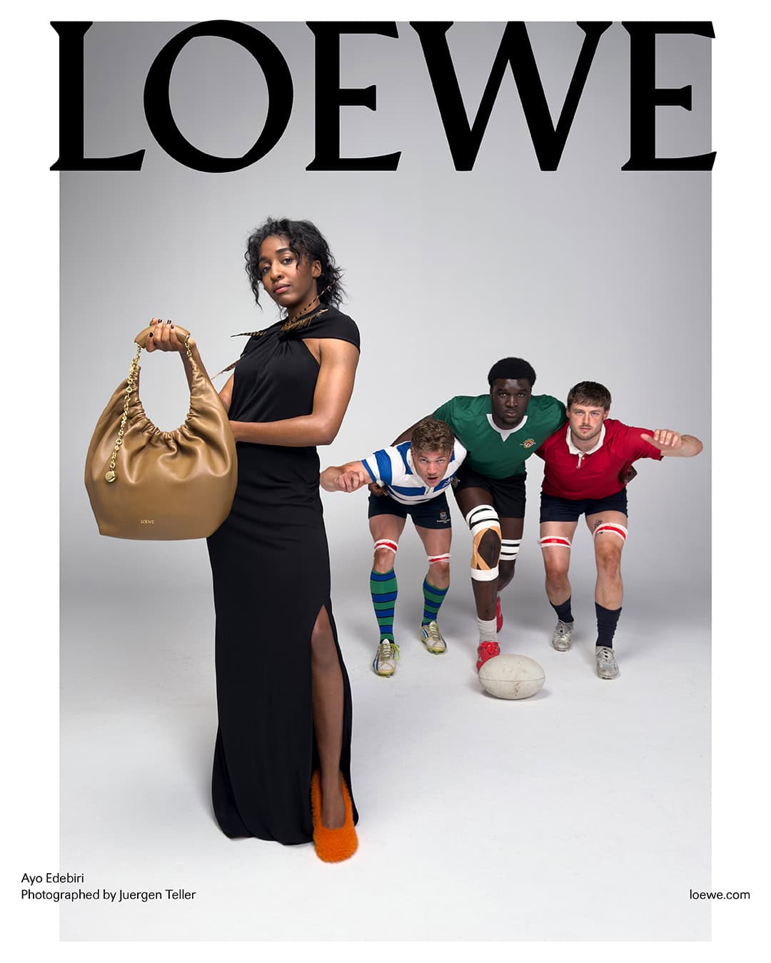 Woman in long black dress holding a tan leather shoulder bag alongside three rugby players against a grey studio backdrop.