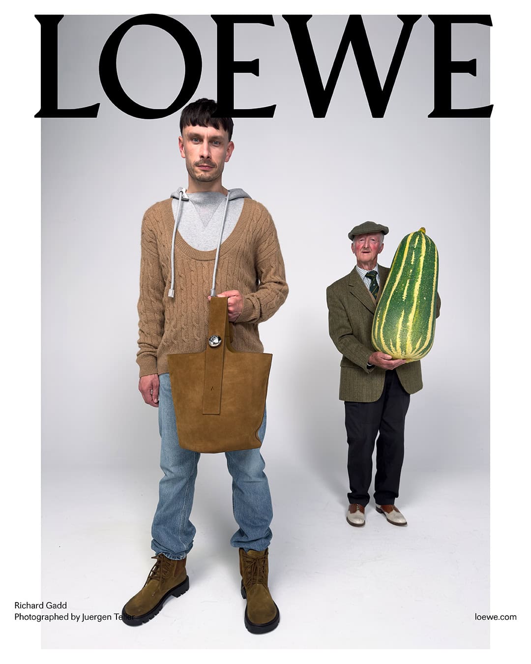 Man standing in beige sweater and blue jeans holding a brown suede bag, with an elderly man holding a giant vegetable in background