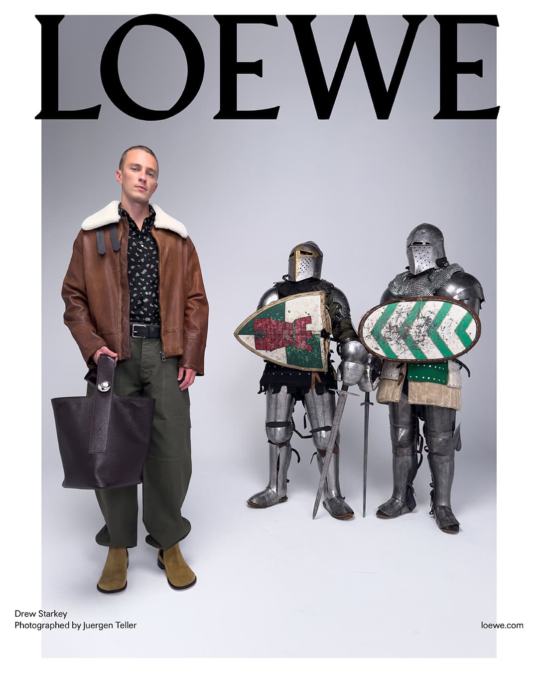 Man wearing a brown shearling collar jacket and dark tote bag standing beside two medieval knights in armor.
