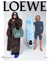 Woman in brown fur coat holding green handbag standing beside a man in a wetsuit holding a surfboard against white background