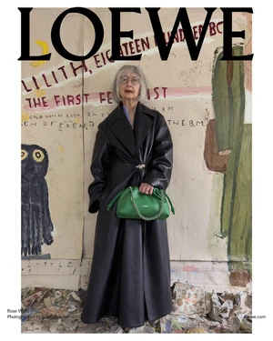 Elderly woman wearing a long dark leather coat and holding a green handbag in front of a painted backdrop