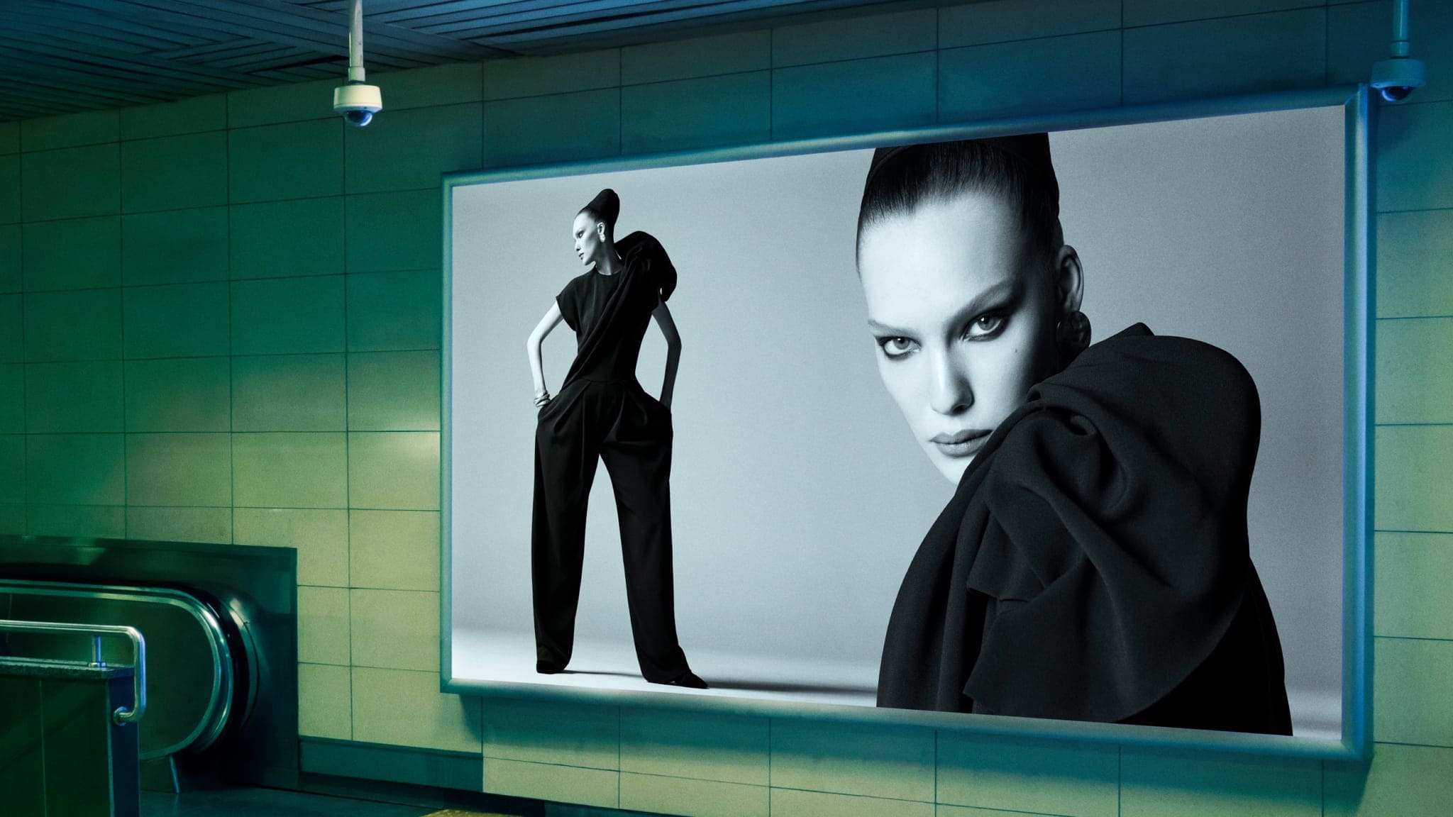 A large fashion billboard displaying black and white portraits of a model in a black outfit inside a green tiled subway station.