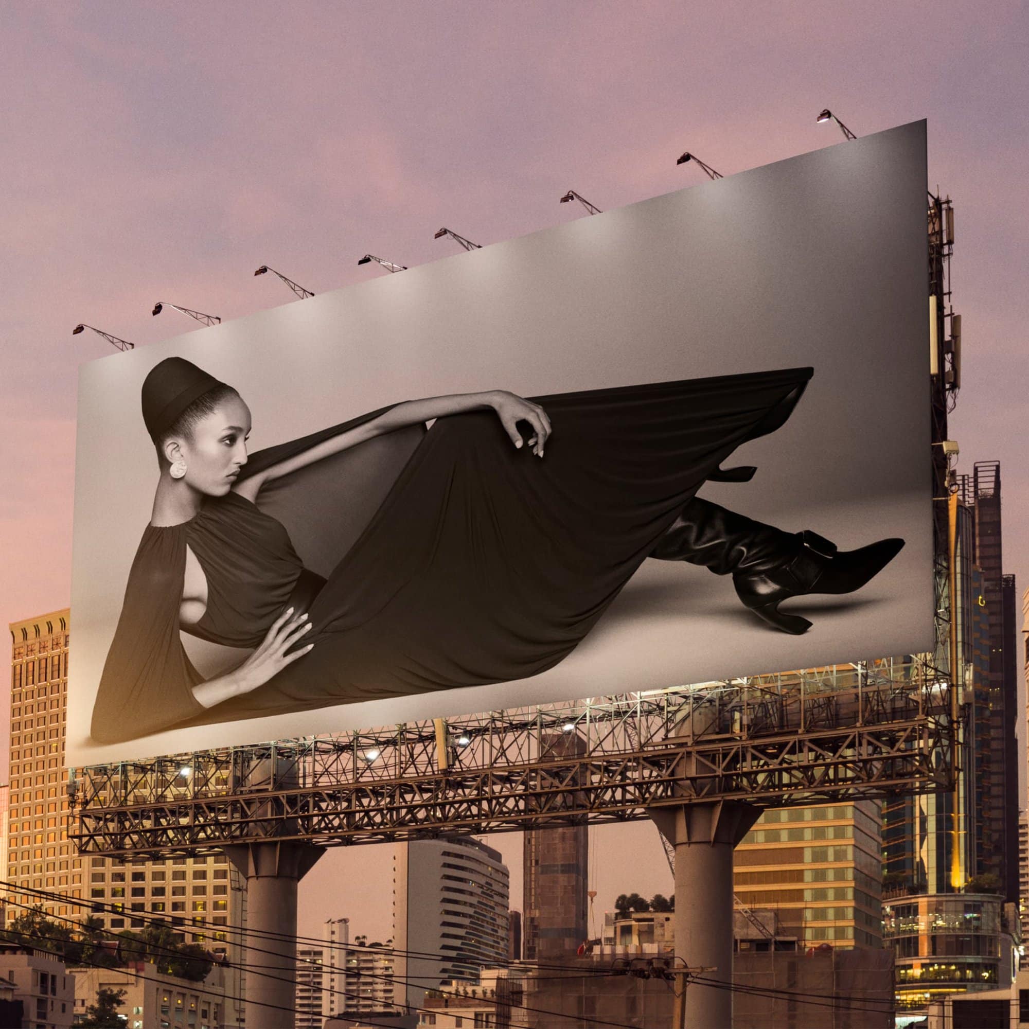 A large urban billboard displaying a black and white fashion advertisement of a model lying down against a sunset cityscape.