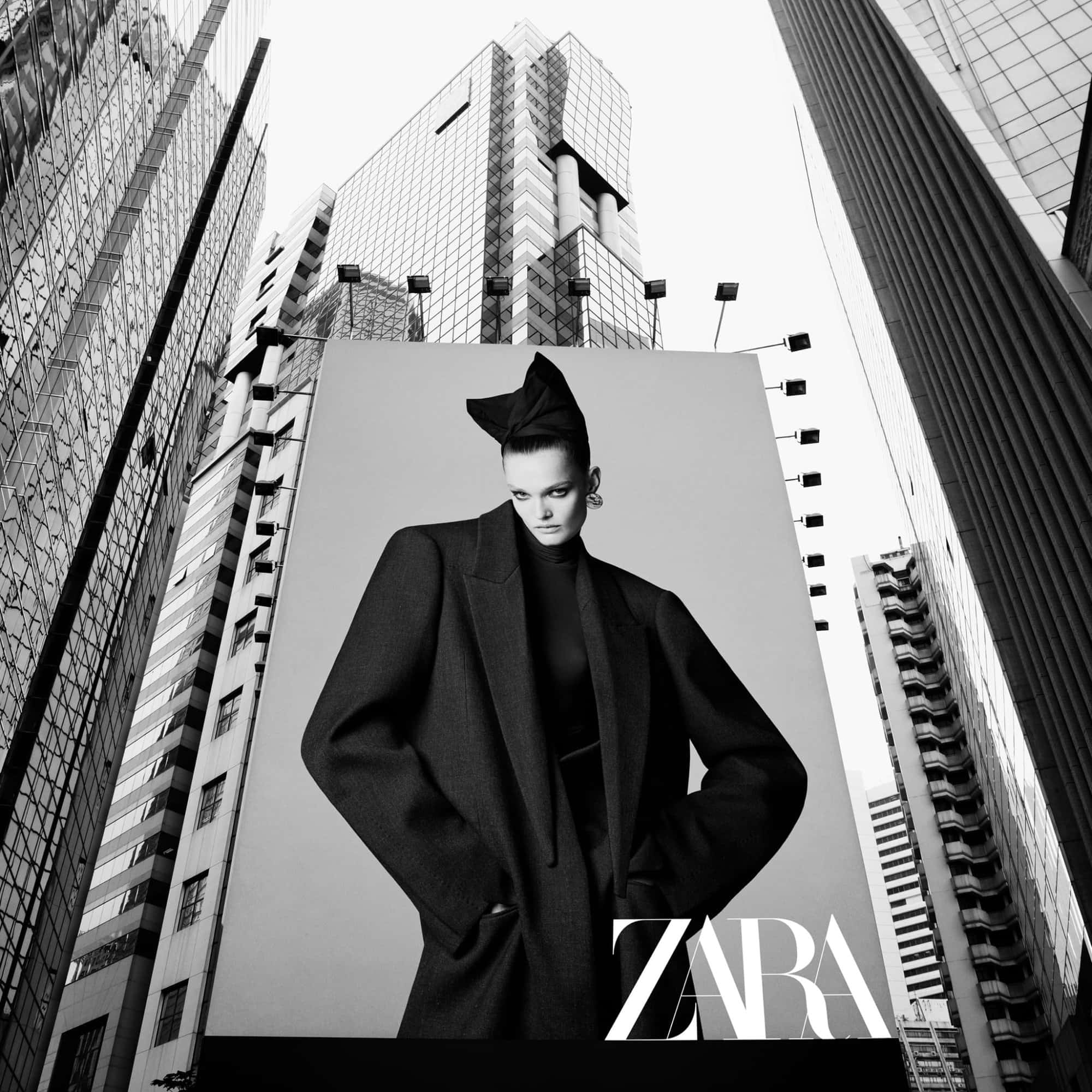 Monochrome low-angle shot of a towering billboard featuring a model in a structured black coat framed by high-rise city buildings.
