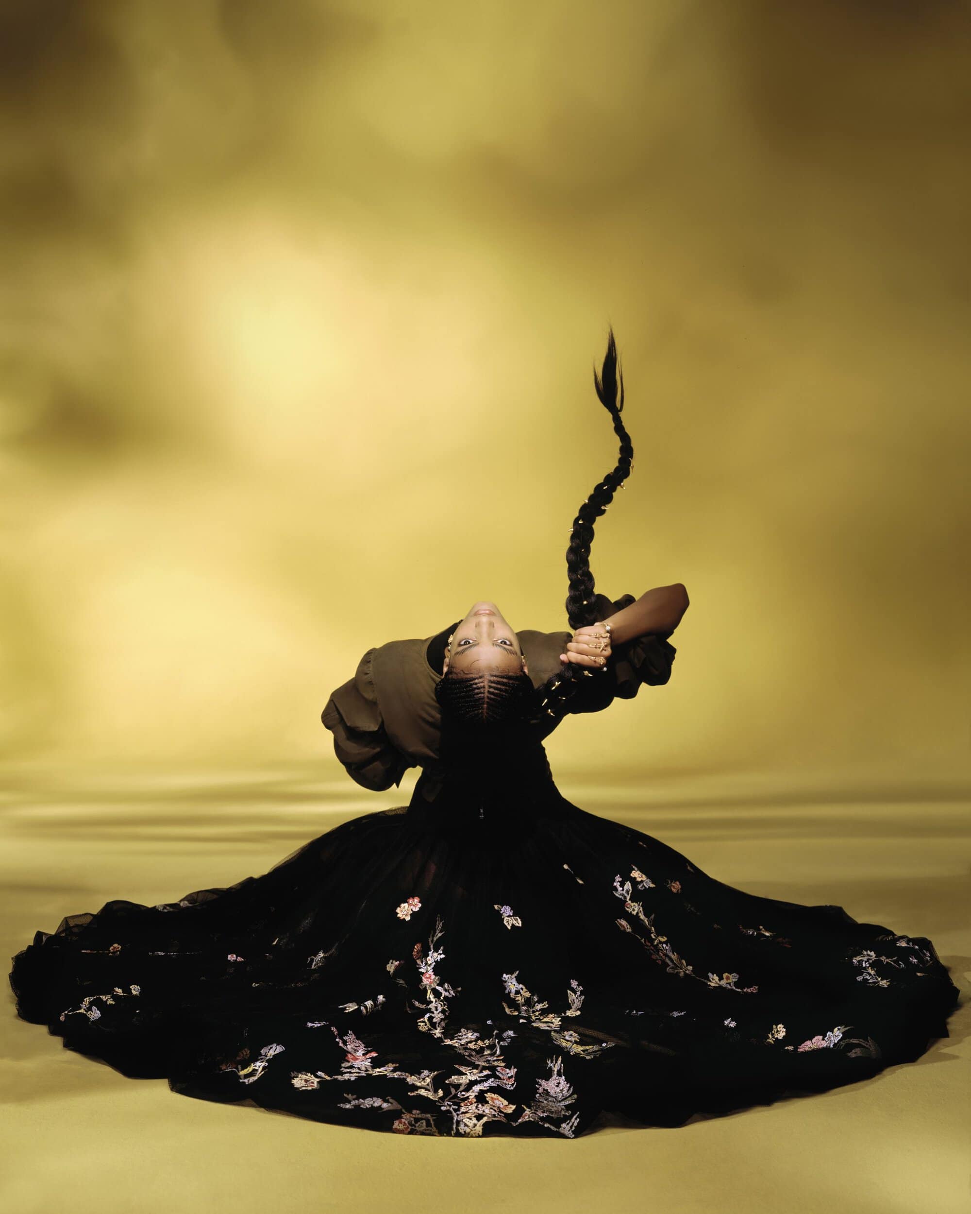 Model in a black embroidered gown with an elaborate braided hairstyle against a golden background